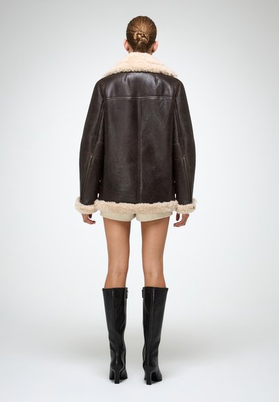 Giacca in shearling marrone con colletto e orlo beige soffice, con vestibilità rilassata e cerniere laterali, indossata sopra pantaloni corti beige e stivali neri.