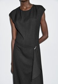 Schwarzes, tailliertes Kleid mit strukturierten Kapuzenärmeln, strukturiertem Stoff und einer asymmetrischen Vorderpartie, die mit einem silbernen Akzentdetail befestigt ist.