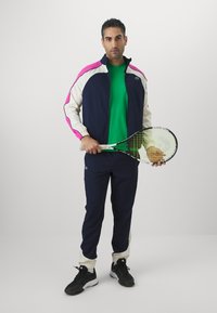 Marineblå og hvit treningsdrakt med rosa detaljer, grønn skjorte og svarte joggesko; holder en tennisracket med grovt grep.