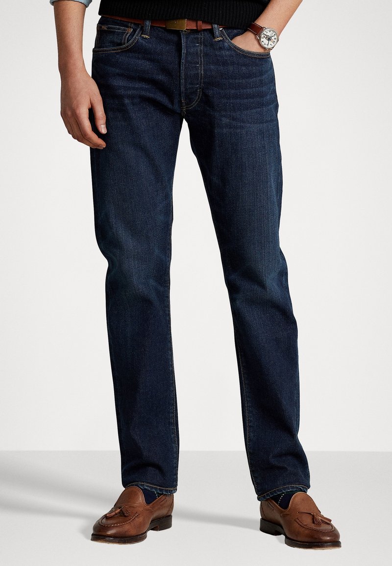 Man som bär mörkblå jeans, brunt läderbälte, bruna tofflor med tofs, en svart tröja och en silverfärgad klocka på vänster handled.