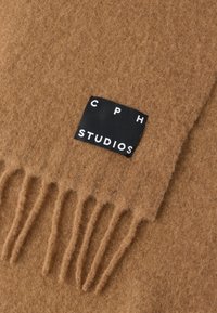 Brązowy filcowy szalik z frędzlami na krawędziach oraz czarną metką z białym napisem "C P H STUDIOS." Miękka tekstura, ciepły materiał.