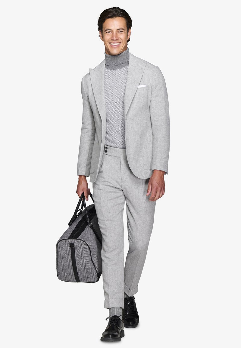 Mand i lys grå dragt med højhalset sweater, der bærer en grå og sort duffelbag, smilende og gående fremad på hvid baggrund.