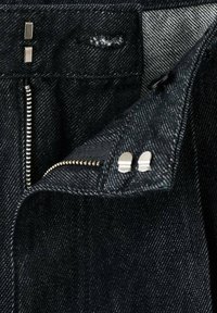 La tela de mezclilla azul oscuro presenta una bragueta con cremallera y hardware plateado, costuras en contraste y dos pequeños acentos metálicos cerca de la cremallera.