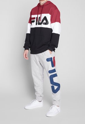 Persona che indossa una felpa con cappuccio FILA rossa, bianca e nera e pantaloni della tuta FILA grigio chiaro con scarpe da ginnastica bianche su uno sfondo grigio uniforme.