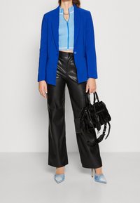 Blå blazer över en randig crop top, kombinerad med vida byxor i svart läder. Accessoarer inkluderar en svart handväska och blå spetsiga skor.