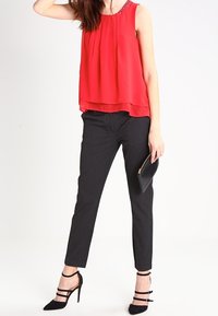 Top rosso senza maniche con un design a strati, collo decorato; abbinato a pantaloni neri su misura e tacchi neri con cinturini.