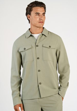 Mężczyzna ubrany w jasnozieloną koszulę na guziki z kieszeniami na piersi oraz dopasowane spodnie, patrzący na bok z ręką w kieszeni.