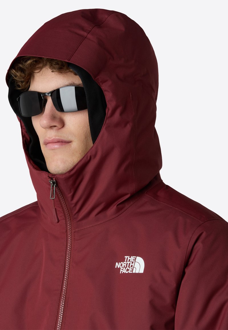 Giacca impermeabile bordeaux con cappuccio, chiusura con zip e logo. Presenta una texture liscia e una vestibilità aderente attorno al viso, abbinata a occhiali da sole scuri.