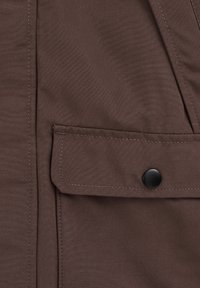 Rjava tkaninska žepna torbica s poklopcem in črnim pritisnim gumbom na zašitem oblačilu, posnetek od blizu, ki prikazuje teksturo in podrobnosti šivov.