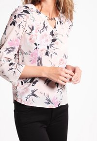 Blusa floreale con base rosa chiaro, motivi di fiori a cinque petali in rosa e nero, e scollatura a oblò con dettagli in catena.