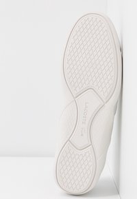 Semelle de basket blanche Lacoste avec un motif de crampons à croisillons et le nom de la marque ainsi que le logo en relief au centre, positionnée verticalement contre un coin blanc.