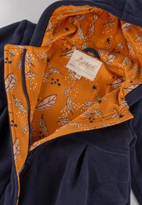 Giacca in pile blu con una fodera arancione decorata con foglie e stelle. Include una cerniera e una chiusura a scatto, con un'etichetta all'interno.