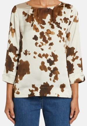 Top crema a maniche lunghe con motivo astratto marrone a macchie, collo tondo e maniche con polsini, indossato con jeans blu.