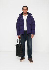 Purple puffer jacket with a hood and quilted design, paired with a white t-shirt, dark jeans, and a black tote bag. Brown shoes.