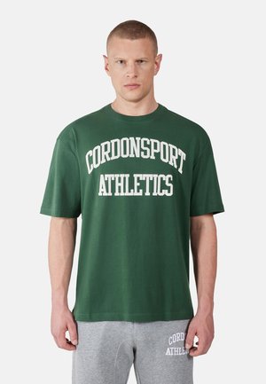 Man wearing dark green "CORDONSPORT ATHLETICS" T-shirt and light gray sweatpants with similar text on left thigh, standing against white background.