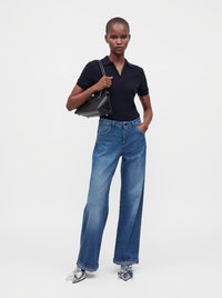 Femme portant un polo noir à manches courtes, jean bleu à jambes larges, chaussures à talons hauts argentées, tenant un sac à main noir sur l'épaule, debout avec une main dans la poche.