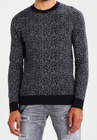 Czarny i szary sweter w wzór jodełki z okrągłym dekoltem i ściągaczami na rękawach, w połączeniu z zniszczonymi jasnymi dżinsami.