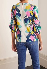 Woman wearing a white blouse with colorful abstract floral print and three-quarter puff sleeves paired with blue jeans, back view.