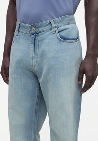 Primer plano de una persona que lleva jeans vaqueros azul claro y una camisa azul metida por dentro, mostrando el bolsillo delantero y las trabillas del cinturón.