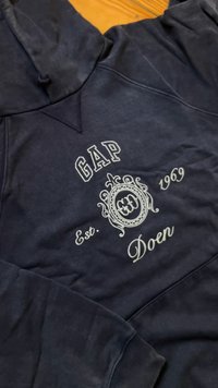 Jūras zila kapuce, kas izgatavota no mīkstas auduma. Iespējo balta izšūta logo "GAP Est. 1969" un "Doen" izsmalcinātā dizainā. Kabata kangurū formā.