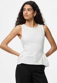 Modella che indossa un top peplum bianco senza maniche e pantaloni neri, in posa con una mano sui fianchi e un'espressione pensierosa.