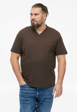 Mann mit Bart und zurückgebundenen Haaren, trägt ein braunes V-Neck-T-Shirt mit kurzen Ärmeln und blaue Jeans, steht mit einer Hand in der Tasche.