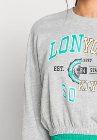 Grå sweatshirt med långa ärmar, kort design, med turkos och svart text samt ett grönt elastiskt midjeband.