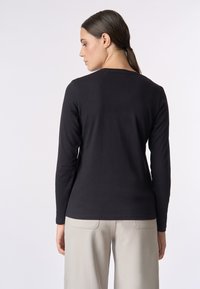 Una donna con i capelli scuri raccolti indossa una maglietta nera aderente a maniche lunghe e pantaloni beige chiari, di spalle contro uno sfondo bianco semplice.