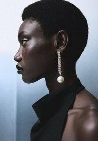 Larga y colgante arete con una perla en la parte inferior, adornada con una cadena brillante de piedras transparentes. El modelo lleva una prenda negra de cuello alto.