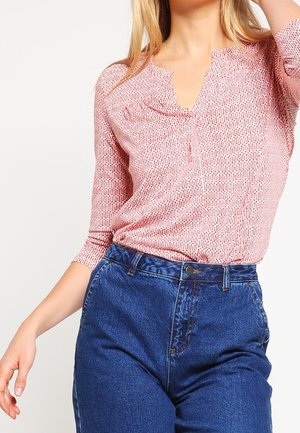 Vrouw draagt een roze blouse met patroon en V-hals en driekwart mouwen, ingestopt in een high-waist blauwe spijkerbroek.