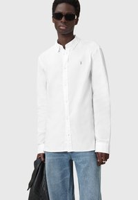 Camicia bianca a maniche lunghe con colletto, caratterizzata da un piccolo logo ricamato sul petto, abbinata a jeans azzurri chiari e occhiali da sole.