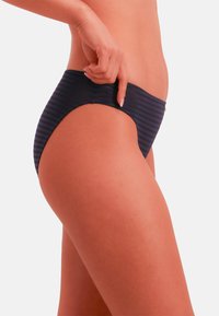 Kvinde iført navyblå bikiniunderdel med striber, der med hånd justerer taljebåndet på hvid baggrund.