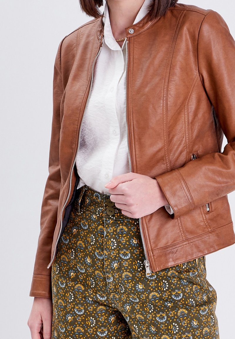 Veste en cuir marron avec fermeture éclair et détails côtelés, portée par-dessus une chemise blanche et un pantalon vert à motif floral.