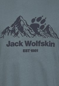 T-shirt en coton bleu présentant un graphique sombre de montagnes et une empreinte de patte. Le texte indique "Jack Wolfskin" et "EST 1981."