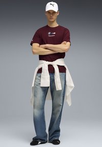 Bordeaux kortærmet skjorte med BMW Motorsport-logo, lyseblå jeans og en hvid skjorte knyttet om taljen. Hvid cap med sort logo.