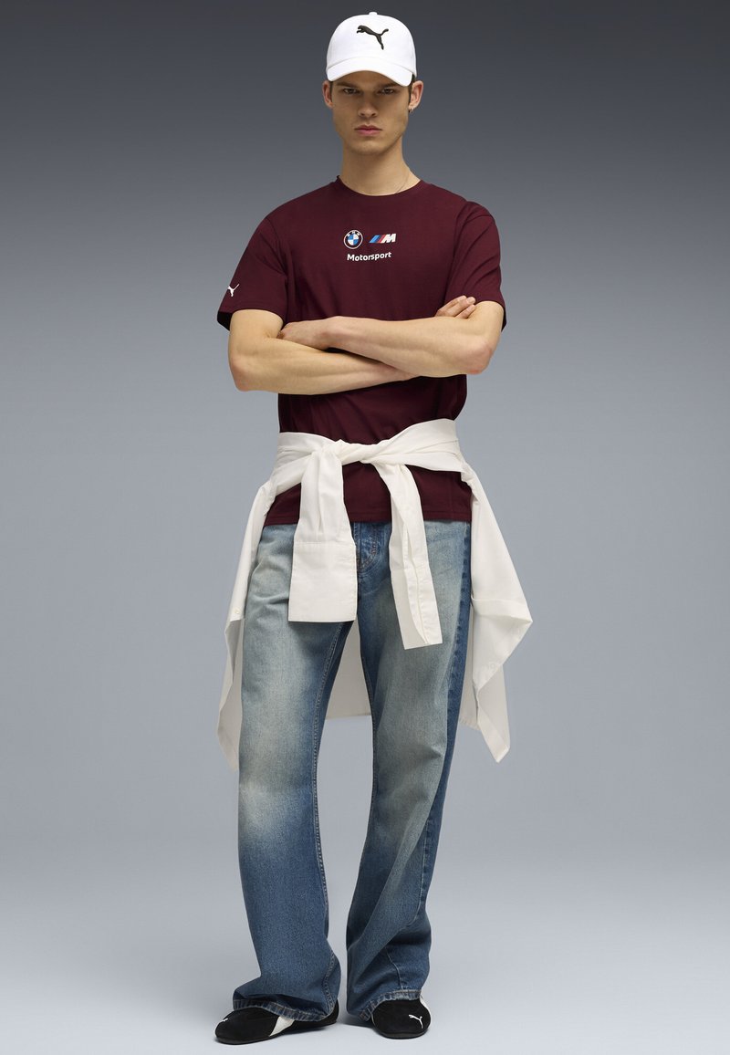 Bordeaux kortærmet skjorte med BMW Motorsport-logo, lyseblå jeans og en hvid skjorte knyttet om taljen. Hvid cap med sort logo.