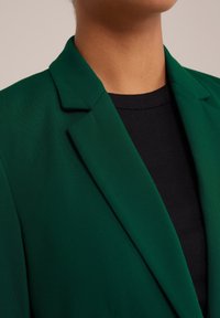 Person som bär en mörkgrön blazer över en svart ribbad topp, som visar halslinjen och övre bröstområdet mot en neutral bakgrund.