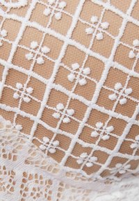 Ljust beige tyg med vit blomsterbroderi i rutmönster. Har spetskant längs botten med intrikata detaljer.