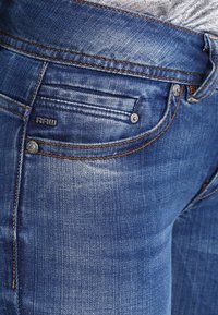 Jeans de mezclilla azul con un corte ajustado, que cuentan con un logo visible en el bolsillo trasero, costuras en contraste y detalles de desgaste mínimos.