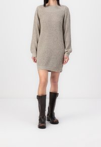 Abito maglione di colore beige chiaro, con maniche lunghe e texture a costine, abbinato a stivali di pelle marrone al ginocchio con fibbie.