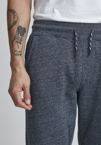 Tumši pelēki sporta bikses ar mīkstu, teksturētu audumu. Funkcijas ietver elastīgu jostasvietu ar baltu auklu un sānu kabatām.