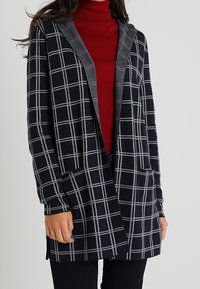 Lång marinblå cardigan med grå rutig mönster, grå huva och framfickor. Bärs över en röd polotröja med långa ärmar och rak skärning.