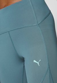 Leggings deportivos en color verde azulado con una textura suave, que presentan costuras planas y un logo de Puma en azul claro en el lado inferior.