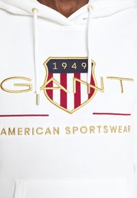 Hoodie bianco con un noto emblema ricamato a forma di scudo, strisce rosse e blu navy, e dettagli dorati, con la scritta "GANT AMERICAN SPORTSWEAR".