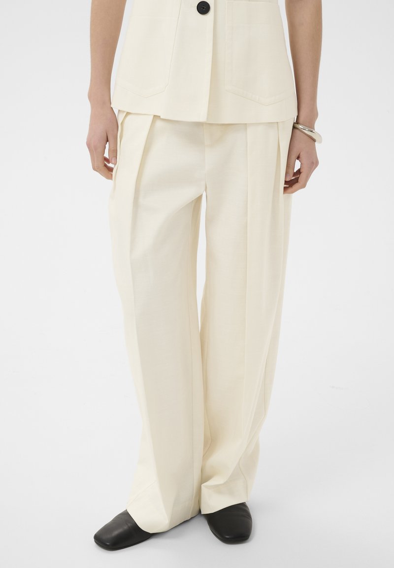 Pantalon large couleur crème associé à un gilet assorti et des chaussures noires à bout carré, porté par une personne debout devant un fond blanc.