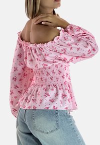 Rosa schulterfreie Bluse mit Blumenmuster, gerafftem Bund und Puffärmeln. Leichter Stoff mit einem Rüschensaum. Kombiniert mit hellblauen Jeans.