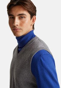 Szary wełniany kamizelka na niebieskim golfie, z dekoltem w kształcie litery V i gładką teksturą. Modelka z krótkimi brązowymi włosami i kolczykiem.