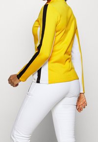 Żółta koszulka sportowa z czarnymi i białymi akcentami, dopasowany krój, długie rękawy i kołnierzyk. W połączeniu z białymi obcisłymi spodniami.