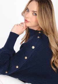 Maglione lavorato a maglia blu navy con un alto colletto, caratterizzato da bottoni dorati lungo una spalla e polsini a coste, che mostra un motivo testurizzato.