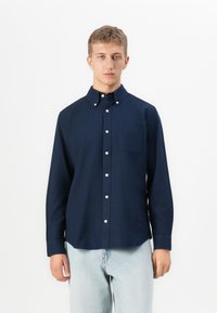 Joven vestido con una camisa de botones azul marino con un bolsillo en el pecho y jeans de color azul claro, de pie frente a un fondo blanco liso.